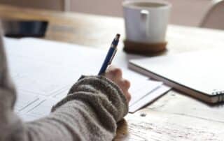 A person writing with a pen on some pieces of paper over a desk with a mug.
