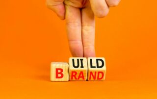 square wood dices that spell Build on one side and Brand on the other. Two fingers show how turning them could reveal one word or the other.