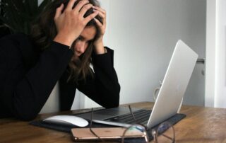 A woman at a desk with a computer and she is holding her head being very overwhelmed.